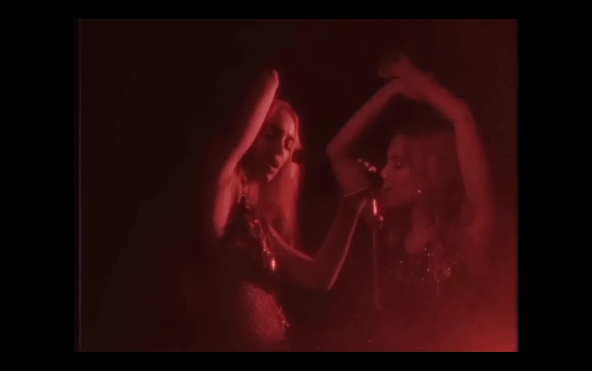 Music video frame of two performers with arms raised in red light