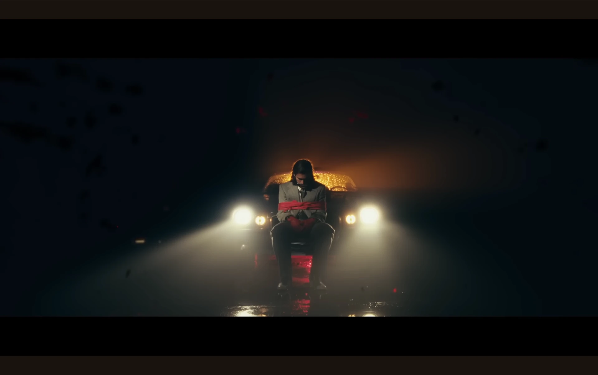 Music video frame of a suited figure tied before a car's headlights